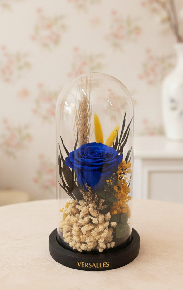 Decorative display with a blue rose and dried flowers under a glass dome, branded 'Versalles'.
