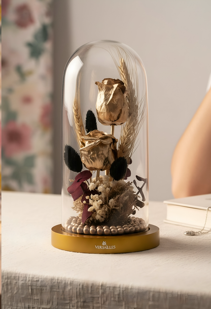 Decorative arrangement of flowers under a glass dome on a table with a floral-patterned curtain in the background.