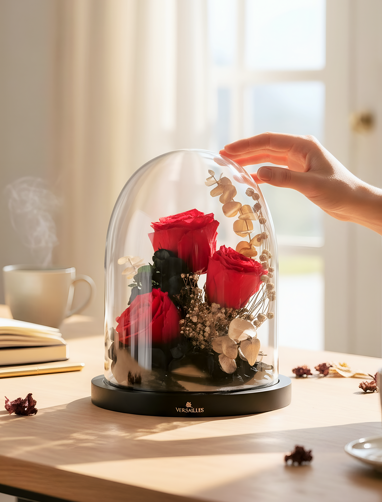 Hand holding a glass dome with red roses and gold leaves on a table.