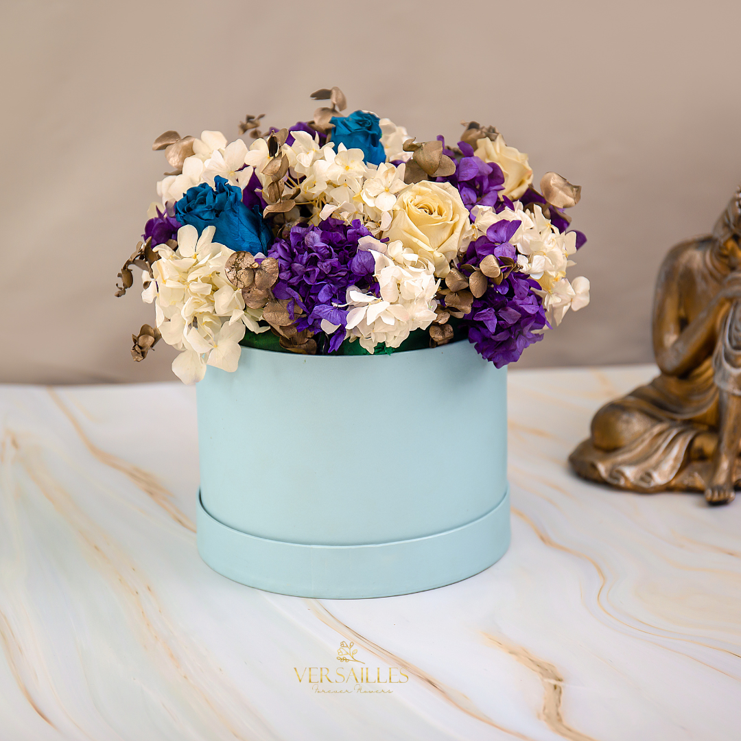 A floral arrangement featuring a combination of purple and white hydrangea and white and blue roses in a light blue container, placed on a marble surface with a golden statue beside it.